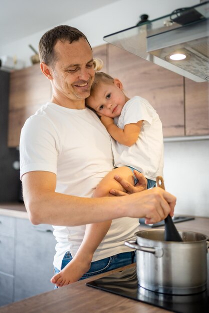 Father with his daughter on his hands cooking soup together in modern home kitchen