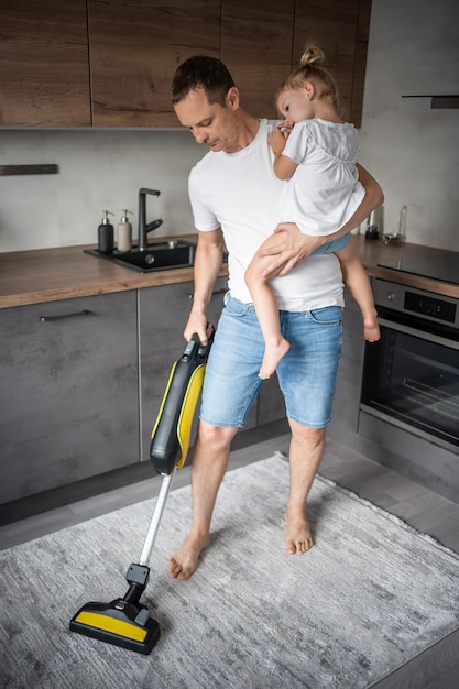 Father with his daughter on his hands vaccuming the floor in the modern home kitchen