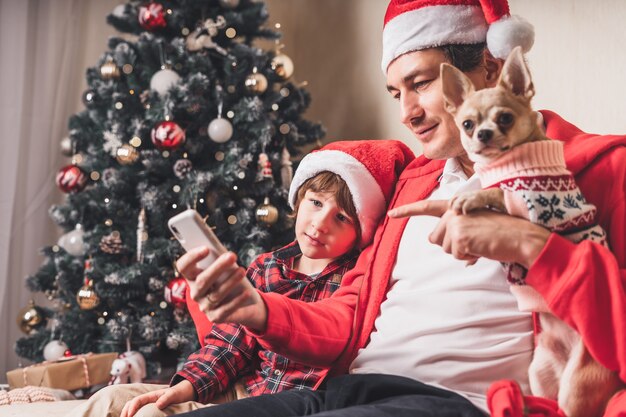 Father with kid and puppy dog in Santa hats holding cell phone