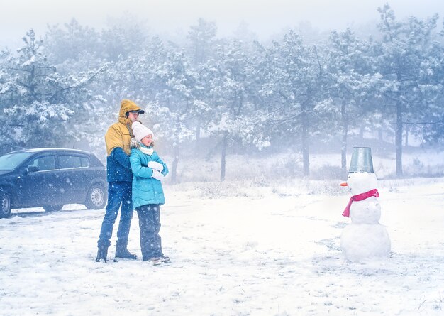 A father with a little daughter in winter clothes are hugging each other in the forest and looking at the snowman.