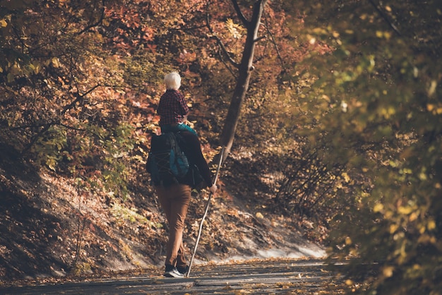 Photo father with son on his shoulders trekking in the autumn forest. back view