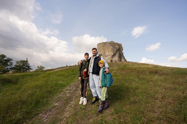 Father with two sons wear backpack hiking near big stone in hill Pidkamin Ukraine