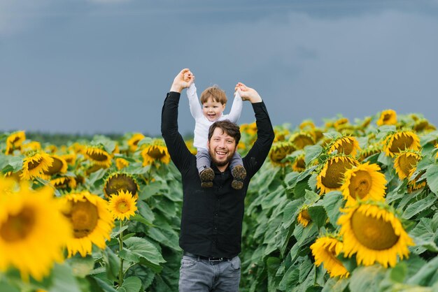 Foto padre con un giovane figlio sulle spalle che cammina e si diverte in un campo di girasoli