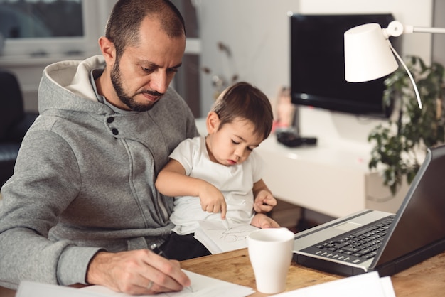father working at home and holding son on his lap