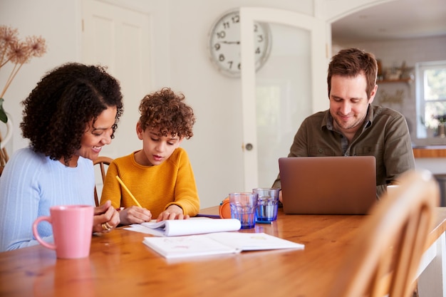 写真 母が台所のテーブルで宿題をする息子を助けるように、父はラップトップで働きます