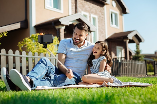 Fatherly love. Pleasant young man sitting on the rug next to his little daughter, hugging her and taking a selfie with a selfie stick while laughing