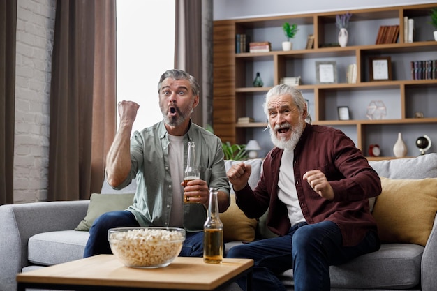 Fathers day adult son with senior father watching sport game on\
tv and celebrating victory sitting on couch at home two generations\
family enjoy spending time together sport and entertainment