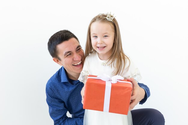 Festa del papà, paternità e concetto di famiglia - il padre abbraccia sua figlia che tiene il contenitore di regalo rosso sopra il muro bianco.