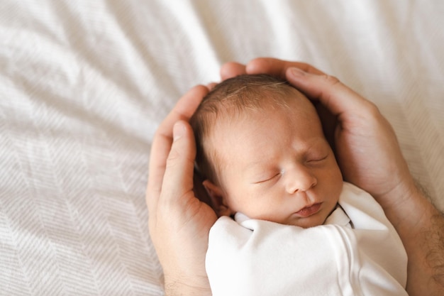 Fatheryoung man holding caucasian hairy brunet cute newborn baby sleepingOne or two week child on bed in male hands indoorsCare love happiness concept Copy space for text