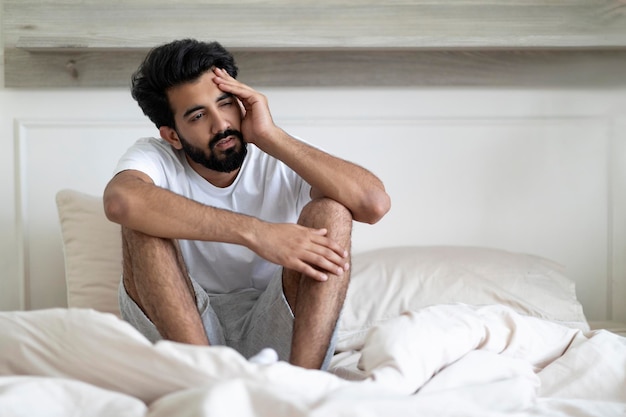 Fatigue concept tired young indian man sitting in bed at home