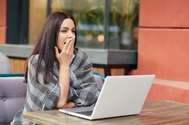 Fatigue overworked female model work at laptop computer for long time, has sleepy expression as yawns, sits against cafe interior