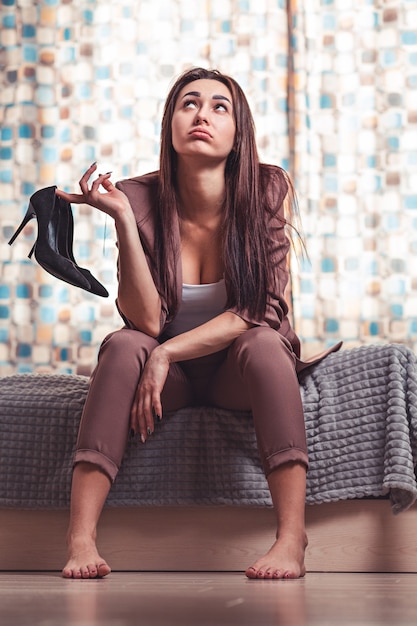 Fatigue and stress after work. A Caucasian woman, a brunette in a suit, with a tired face, is sitting on the bed barefoot and holding shoes in her hands. Window in the background.