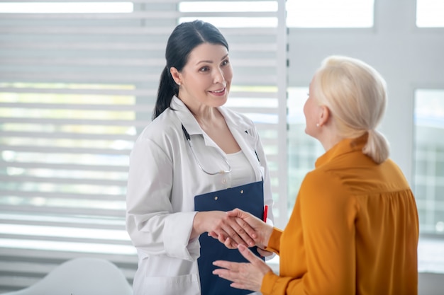 Favorable communication. Satisfied woman in a white coat and a stethoscope with a folder and the patient in a yellow blouse shaking hands.