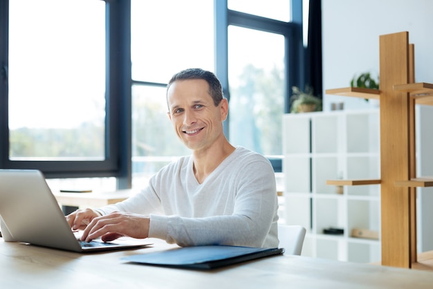 Favoriete baan. Vrolijke positief opgetogen zakenman typen op de laptop en glimlachen terwijl u geniet van zijn baan