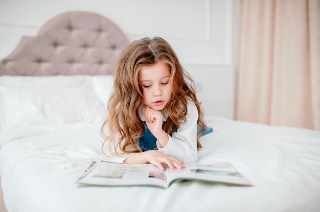 好きなおとぎ話。ベッドの中で女の子は本を読みます。快適なベッドルームでお楽しみください。子供の長い髪はリラックスして本を読みます。