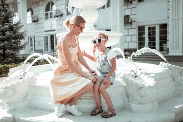 Favorite person. Little girl wearing big sunglasses sitting on the fountain spending time with her loving mother.