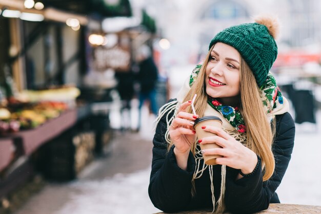 好きな季節。クリスマスマーケットで自由な時間を過ごし、コーヒーを飲みながら、積極性を表現する素晴らしい女の子。喜びの笑顔で目をそらしている女性