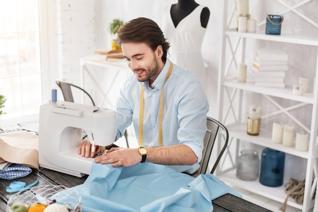 Favourite activity. Exuberant dark-haired tailor smiling and working on a sewing machine