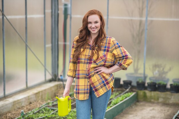 Favourite hobby. Happy young adult woman with long ginger hair with light green watering can standing in greenhouse in optimistic mood