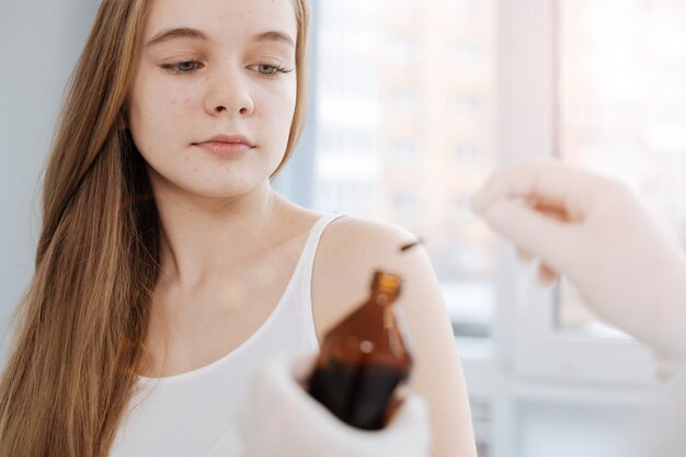 Fearless motionless charming teenager sitting in the hospital and waiting for treatment while dermatologist holding the bottle of iodine and cotton swab