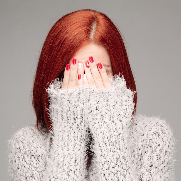 Fed haired unrecognizable woman wearing warm gray seater hiding face by hands.