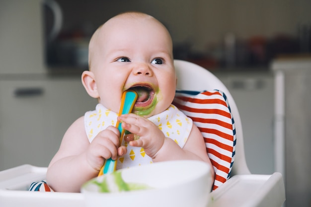 Feeding Messy smiling baby eating with a spoon in high chair Babys first solid food