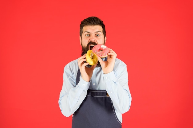Feel hunger bearded man in chef apron brutal waiter on kitchen mature man red background skilled baker with donut dessert hungry hipster cook doughnut temptation while diet male cooking bakery