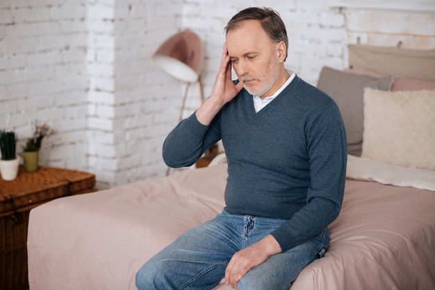 Feeling bad. Old handsome man sitting on bed and touching his temple because of terrible headache.