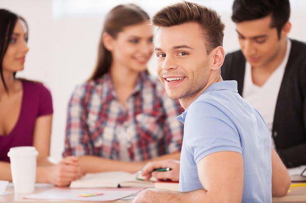 Fiducioso per il suo esame finale. quattro studenti allegri seduti insieme alla scrivania e studiano mentre un uomo si guarda alle spalle e sorride