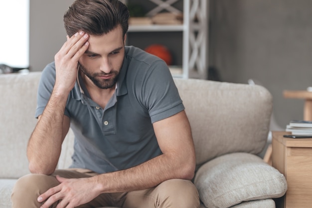 Feeling depressed. Desperate young man keeping his hand on forehead while sitting on the couch at home