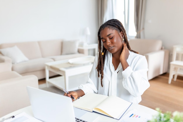 Sentendosi esausta giovane donna africana frustrata che sembra esausta e si massaggia il collo mentre è seduta al suo posto di lavoro