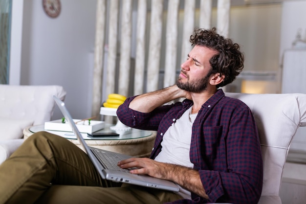 Feeling exhausted. Frustrated young handsome man looking exhausted while sitting at his home and working on his laptop while massaging his neck, Neck pain