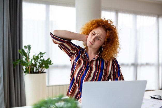 Photo feeling exhausted frustrated young woman looking exhausted and massaging her neck while sitting at her working place