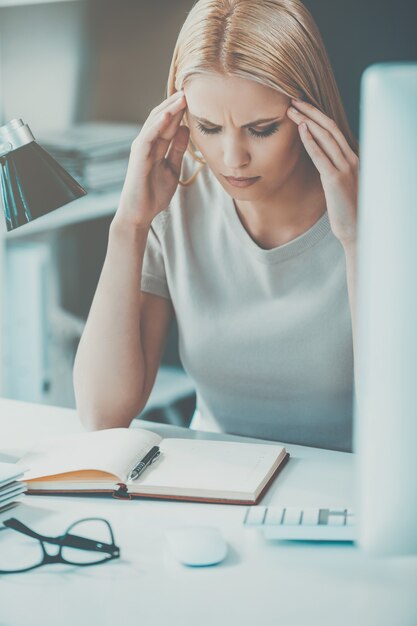 疲れた感じ。欲求不満の若い女性がオフィスの職場に座っている間、頭に触れて目を閉じたまま