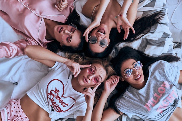 Feeling free to be happy. Top view of four playful young women in pajamas and eyewear smiling while lying on the bed at home