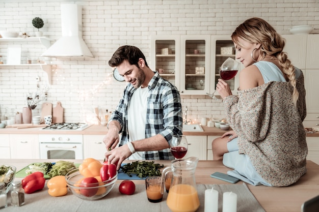 Feeling great. Dark-haired handsome man in a checkered shirt looking contented while cutting vegetables for breakfast