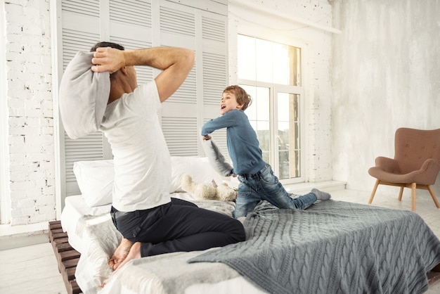 Feeling great. Nice joyful little fair-haired boy smiling and playing with his daddy at home and they having a pillow fight