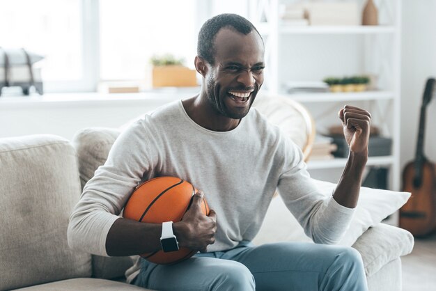 幸せだ。ボールを持って、自宅のソファに座って笑っているハンサムな若いアフリカ人