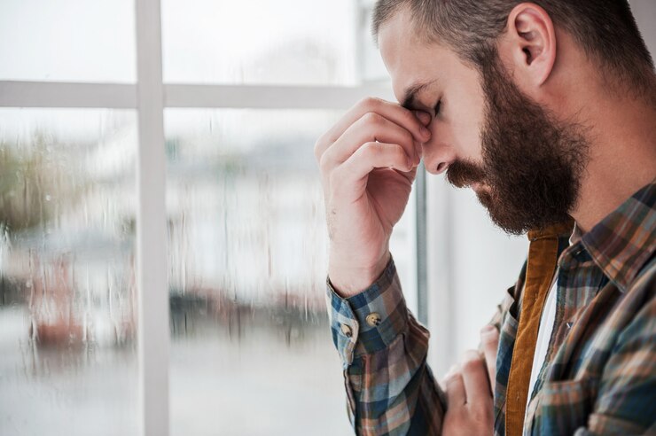 Photo feeling hopeless. depressed young bearded man keeping eyes closed and touching his face while standing near the window