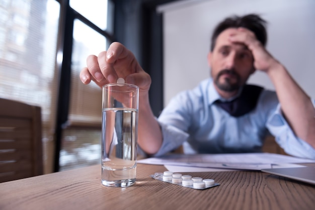 Feeling ill. Exhausted gloomy pessimistic man holding his head and putting a pill into the glass of water while taking medicine