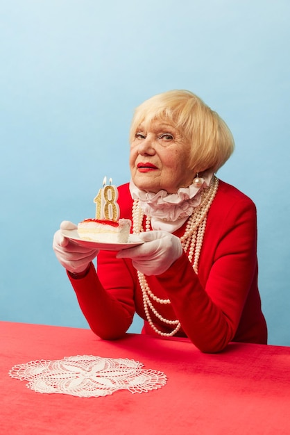 Feeling like 18 Beautiful old woman grandmother in stylish red dress and pearl necklace posing over blue studio background Concept of age fashion and emotions