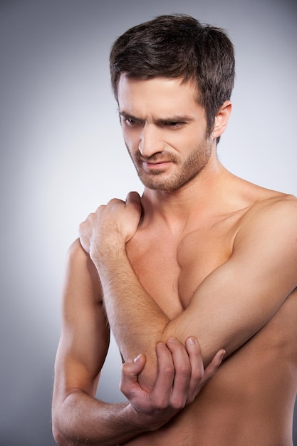 Feeling pain in elbow. Young muscular man touching his elbow and expressing negativity while standing isolated on grey background