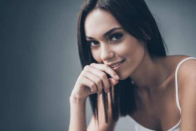 Feeling playful. Attractive young woman holding hand on chin and looking thoughtful while sitting against grey wall