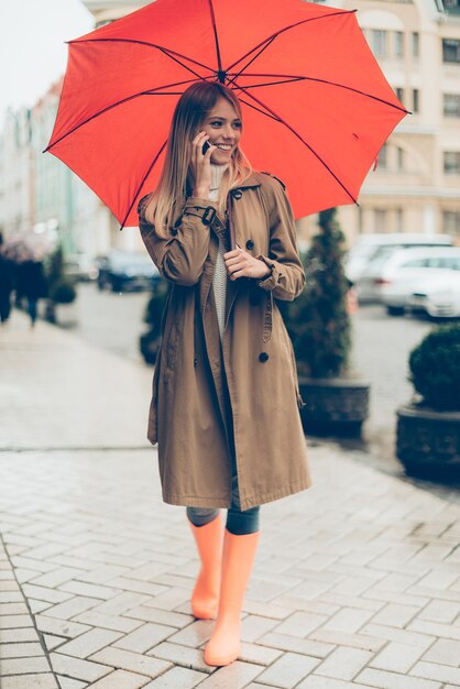 Feeling ptotected at this autumn day. Full length of attractive young smiling woman in colorful rubber boots carrying umbrella and talking on the mobile phone while walking by the street