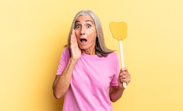 Feeling shocked and scared, looking terrified with open mouth and hands on cheeks and holding a fly swatter