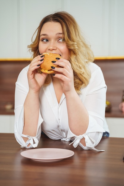 Sentirsi timido. ragazza attraente che si appoggia i gomiti sul tavolo mentre morde il suo hamburger