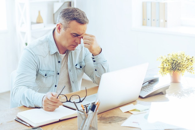 Foto sensazione di nausea e stanchezza. uomo maturo frustrato che sembra esausto mentre è seduto al suo posto di lavoro e porta gli occhiali in mano