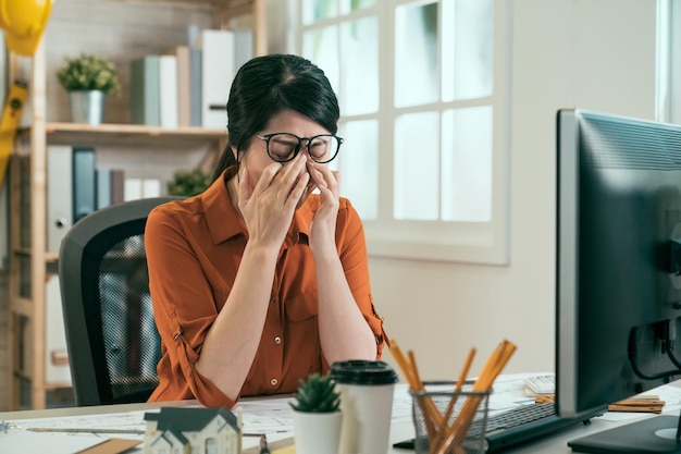 Feeling tired. frustrated young asian woman architect looking\
exhausted and covering face with hands while sitting at working\
place in architecture company. girl engineer rubbing hurt eyes in\
office