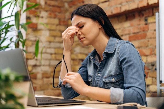 Photo feeling tired and stressed. frustrated young woman keeping eyes closed and massaging nose while sitting at her working place in office