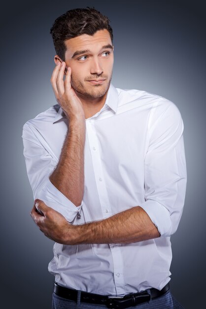 Feeling uncertain. Handsome young man in white shirt holding hand in hair and looking away while standing against grey background